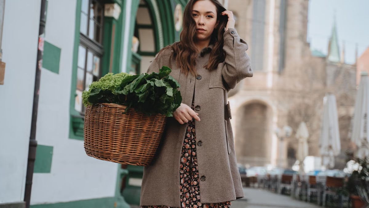 A woman in a brown coat carries a wicker basket of green vegetables while strolling through Erfurt, Germany.